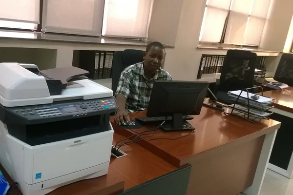 Mwanamume akifanya kazi kwenye kompyuta karibu na mashine ya kuchapisha katika ofisi nchini Tanzania.